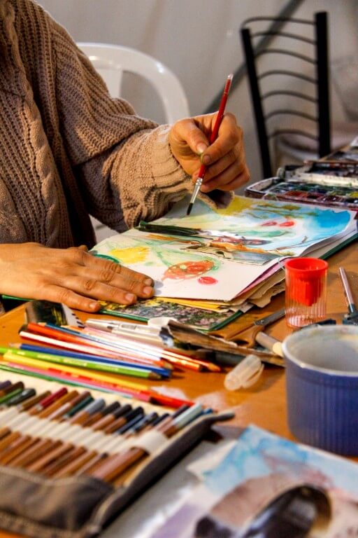 איזה מציירת בסטודיו עם מכחול על מחברת, חומרי אומנות וצבעים מסביב